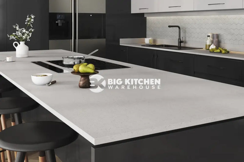 Modern kitchen with a large light gray island featuring Vis Oya Square Edge Laminate Kitchen Worktops, black cabinets, built-in stove, fruit bowl, flower vase, and stools. The background shows a sink, backsplash, refrigerator, and stylish countertops.